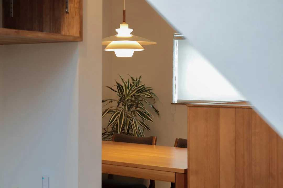 階段下のヌックから見たダイニング。ダイニングテーブルの上を照らす北欧デザインのペンダントライト。奥には観葉植物と窓からの光が見える