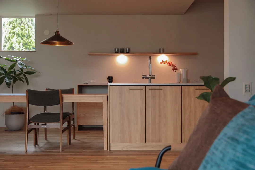 間接照明が埋め込まれた造作棚と、木のキッチン、ダイニングの一部を雰囲気良く写している。手前にはソファに置かれたクッションが見える。