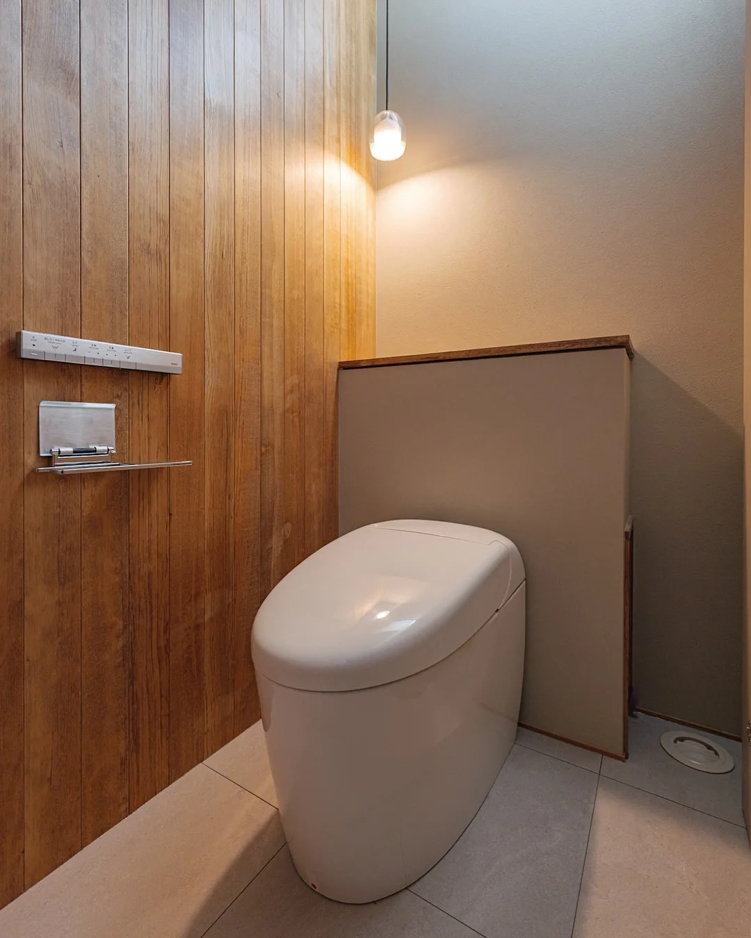 タンクレスタイプの便器と、一面に配された木目の壁、小さなペンダントライト。暖かい雰囲気。