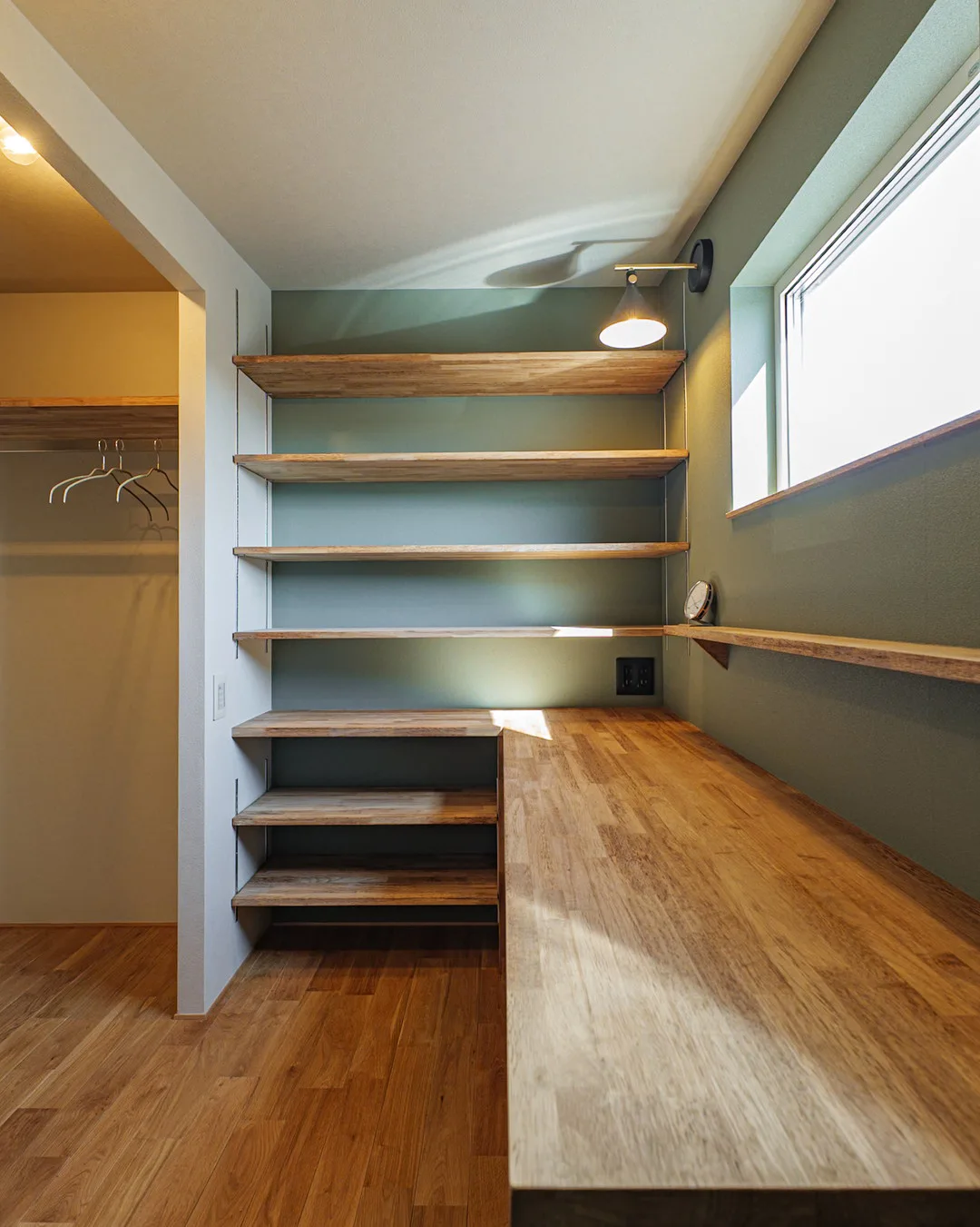 小さい書斎。壁一面に設置された長い木製カウンターと可動棚、手元を照らす黒いブラケットライト。