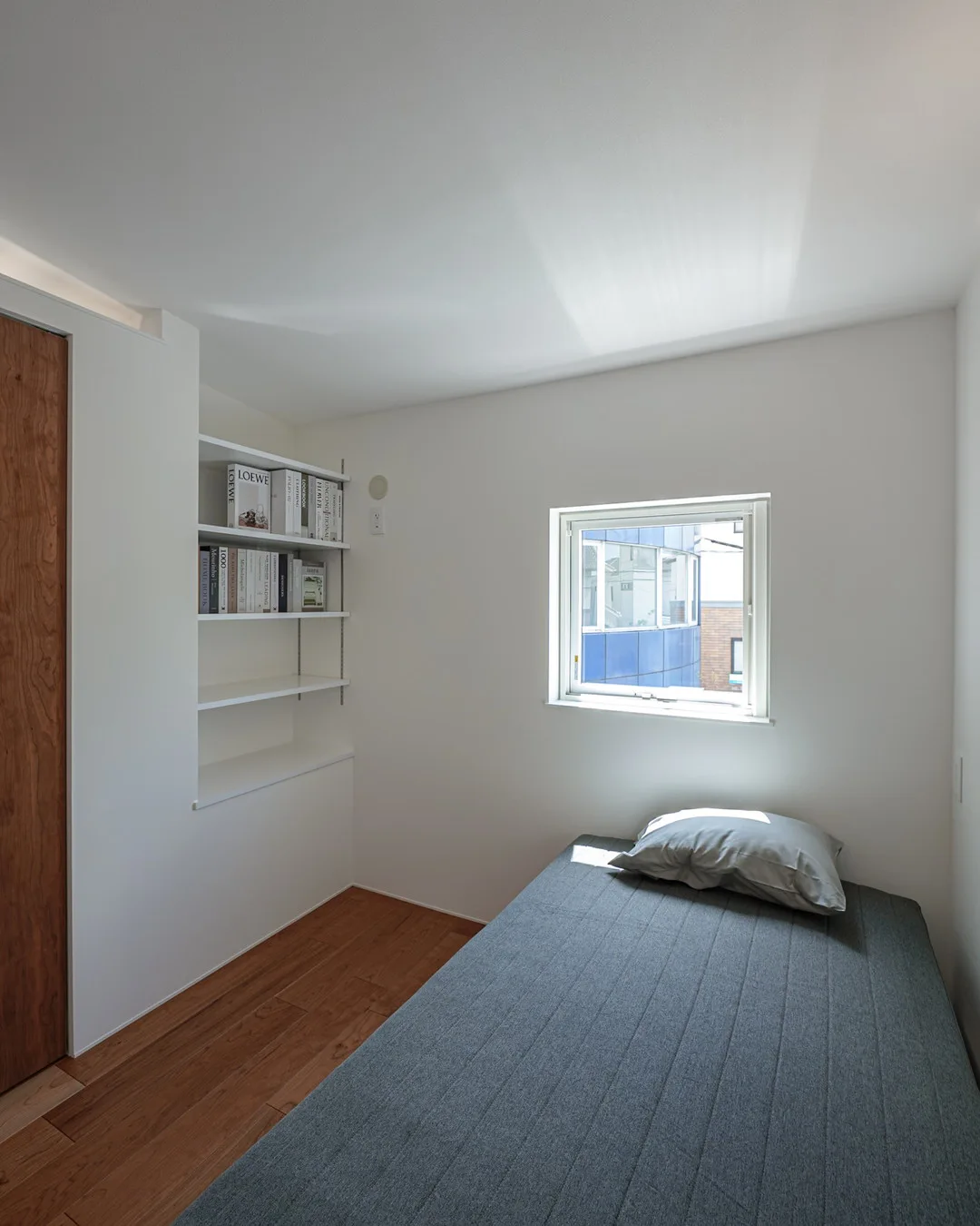 小さな正方形の窓から光が入る部屋。ベッドと壁埋め込みの本棚が備え付けられている