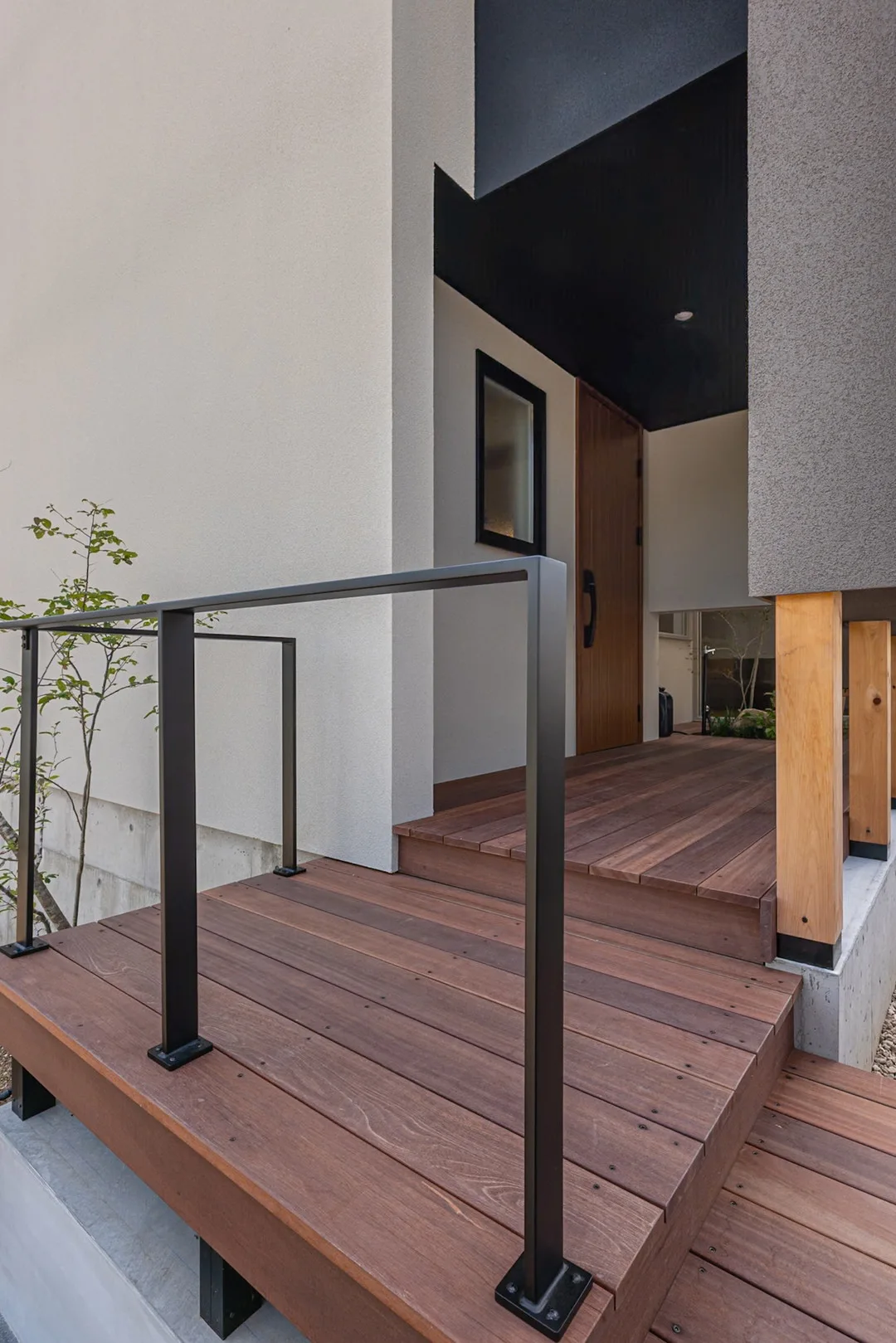 木目調の玄関ドアへと続くウッドデッキの階段と、シンプルな黒い手すり